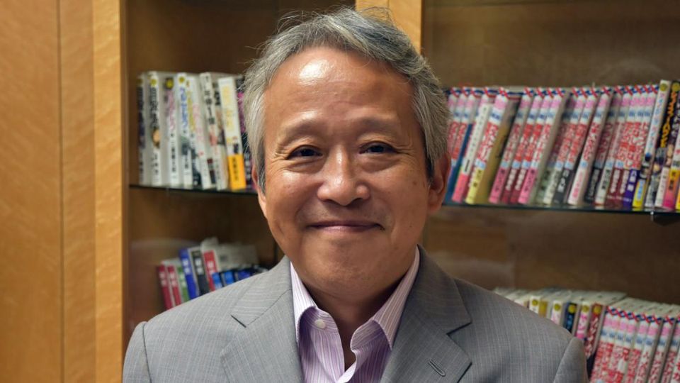 Foto de Kazuhiko Torishima sorrindo, de pé, em frente a uma estante coberta por mangás.